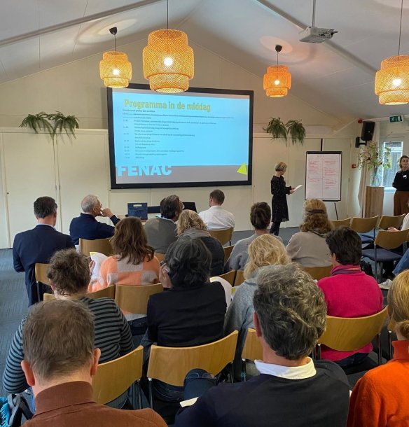 Zaal met mensen en presentatie op een scherm tijdens een ledenbijeenkomst